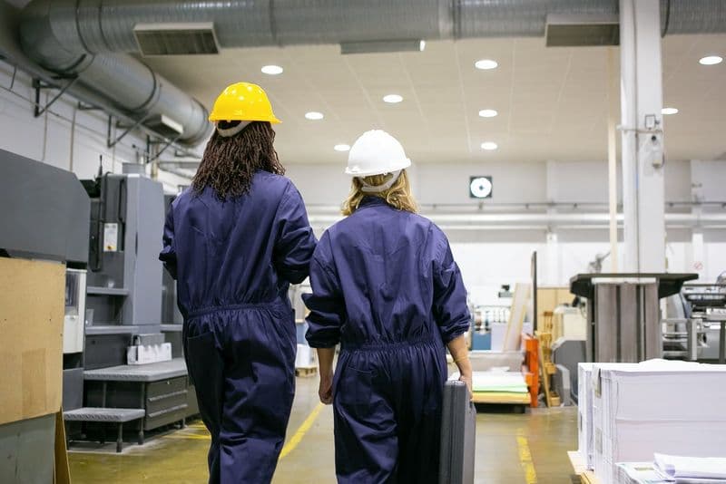 Trabalhadoras de fábrica com capacetes caminhando pelo chão de produção da indústria